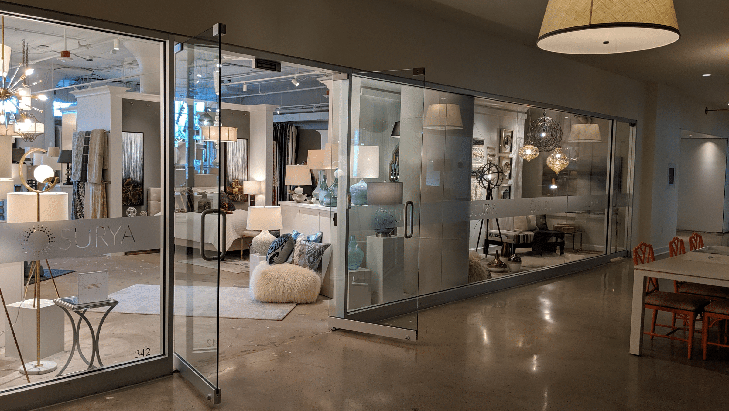 Glass-walled showroom displays home decor items including lamps, rugs, cushions, and furniture, with "SURYA" branding on the glass doors.