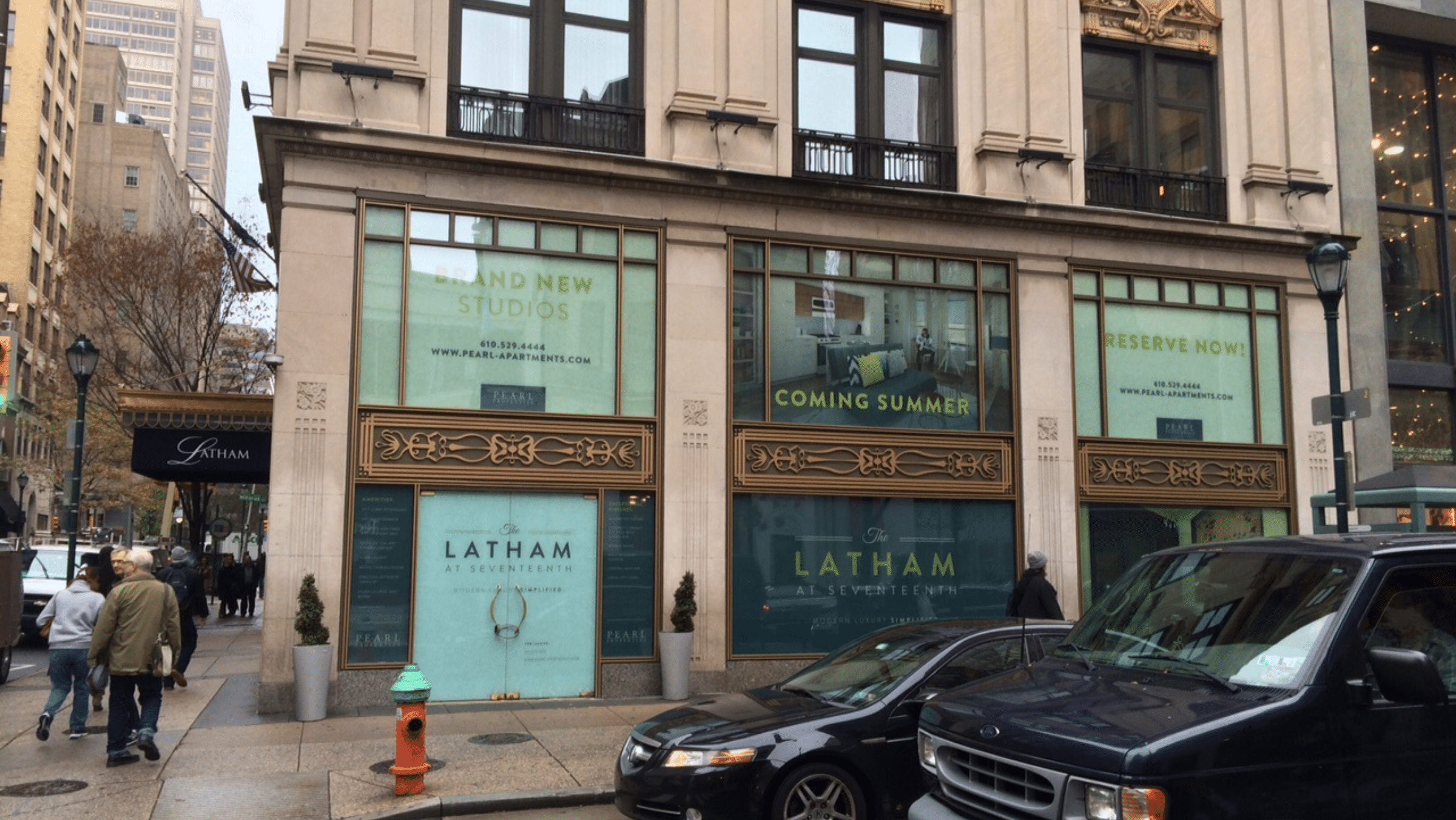 Street view of a building with large signs advertising new studios at The Latham; vehicles and pedestrians are visible in front.