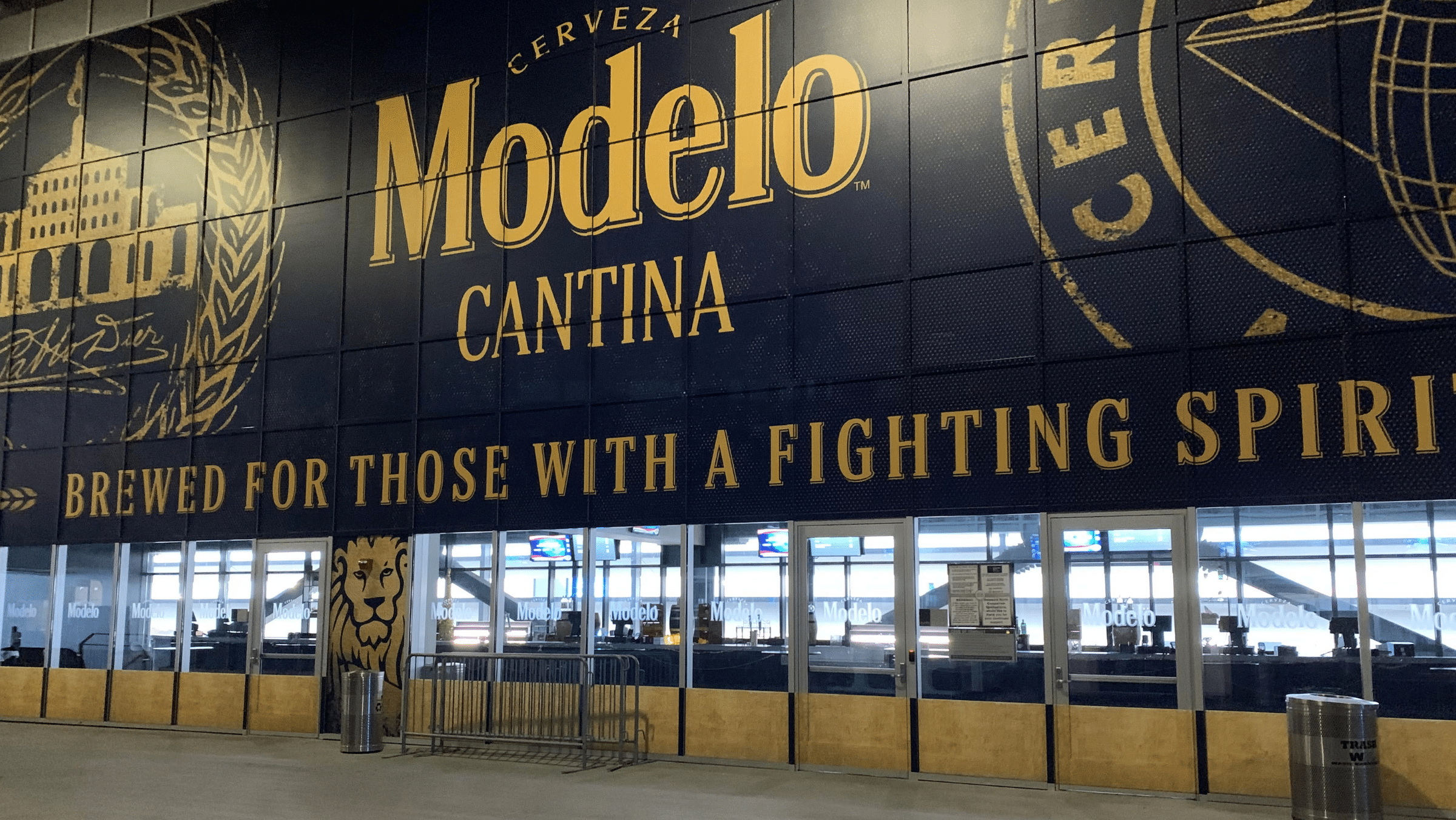 Large Modelo Cantina sign with the slogan "Brewed for those with a fighting spirit" displayed above ticket windows and barriers at an indoor venue.