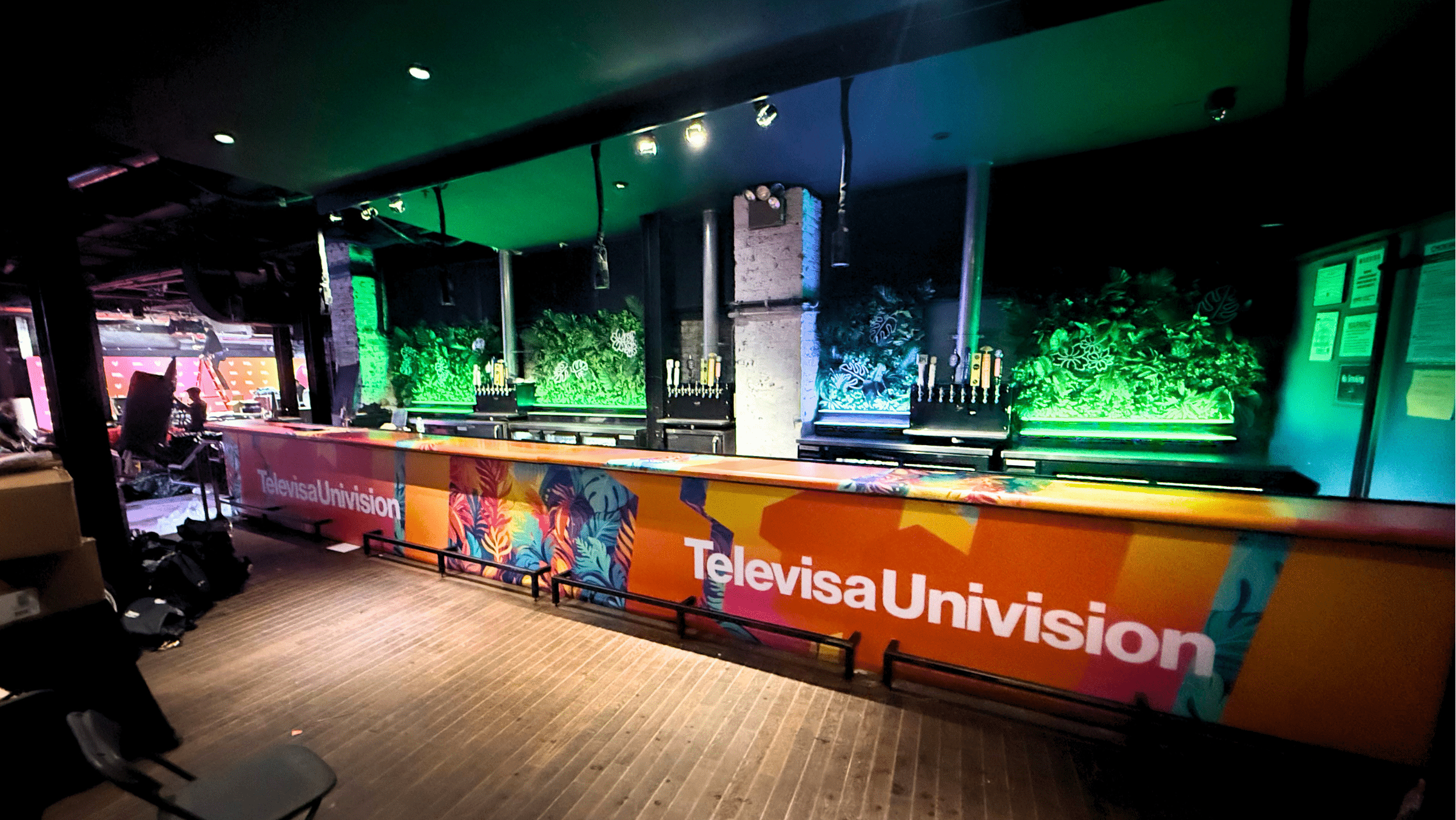 A long bar counter with "TelevisaUnivision" branding, colorful graphics, and beer taps, set against a green plant wall in a dimly lit indoor space.