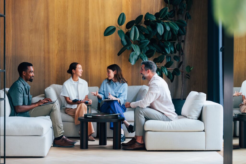 Four people sit on sofas in a modern lounge area, engaged in conversation around a low black table, with a large plant in the background.